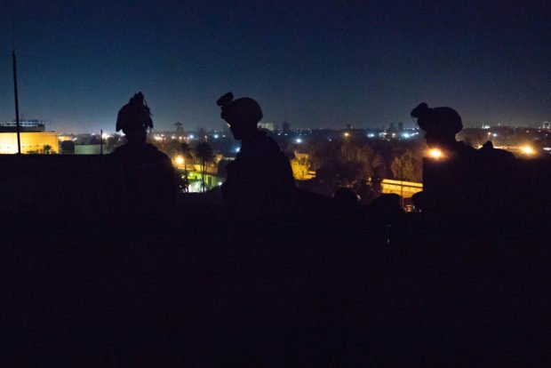 Three Marines wearing combat equipment are silhouetted against a city skyline at night.