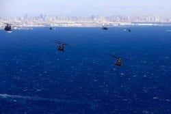 A group of military helicopters fly above the water with a coastline behind them.