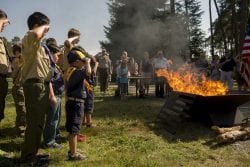 Several boys in Boy Scout uniforms salute a blazing fire pit as adults do so in the background.