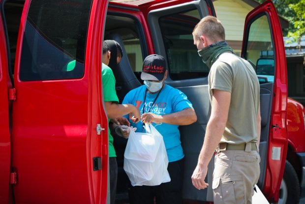 Two soldiers wearing face masks and civilian clothes flank a woman as she receives a white plastic bag containing meals.
