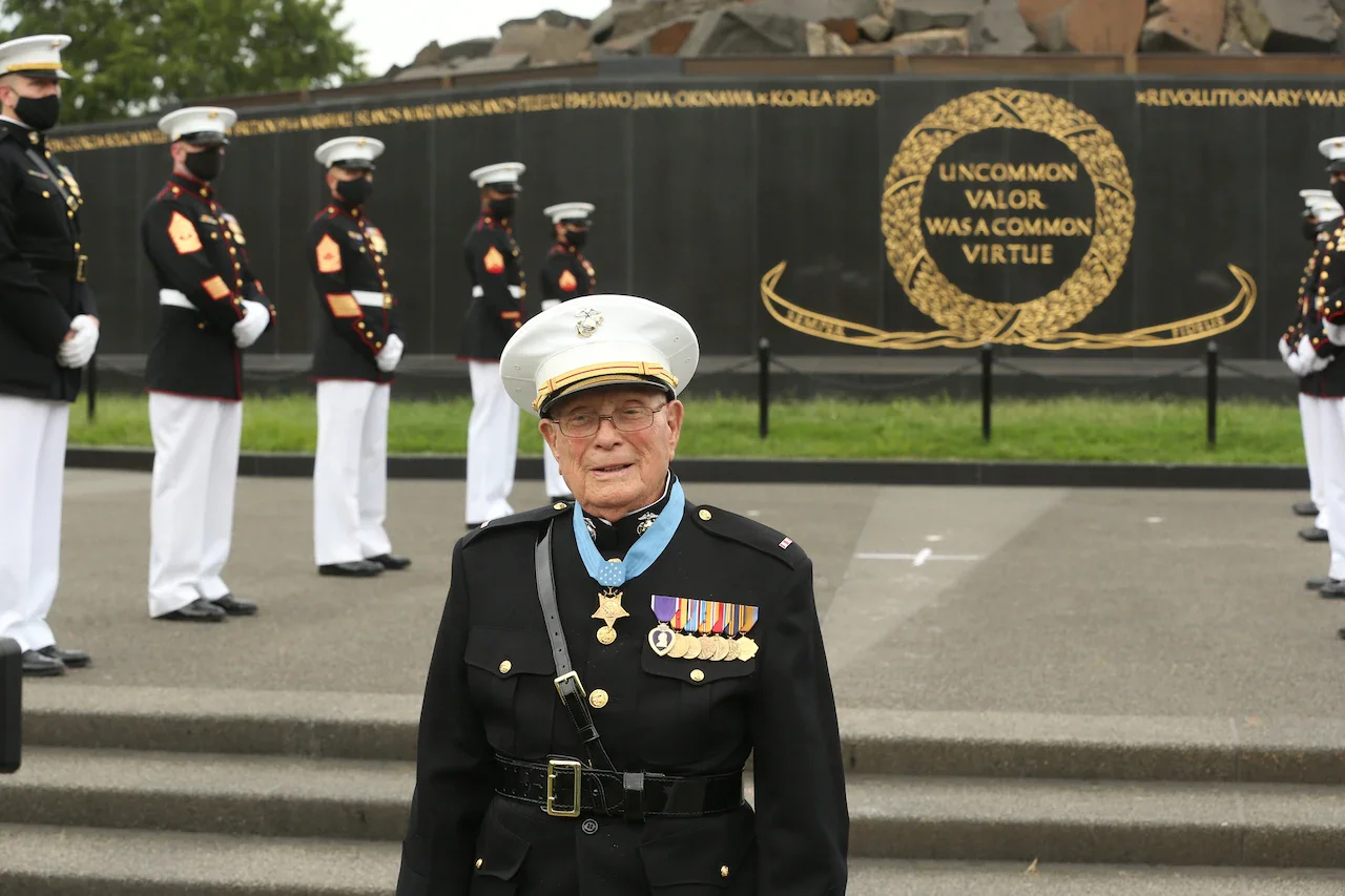 A Marine Corps veteran in uniform and wearing the Medal of Honor poses for a photo. Marines stand in formation behind him.