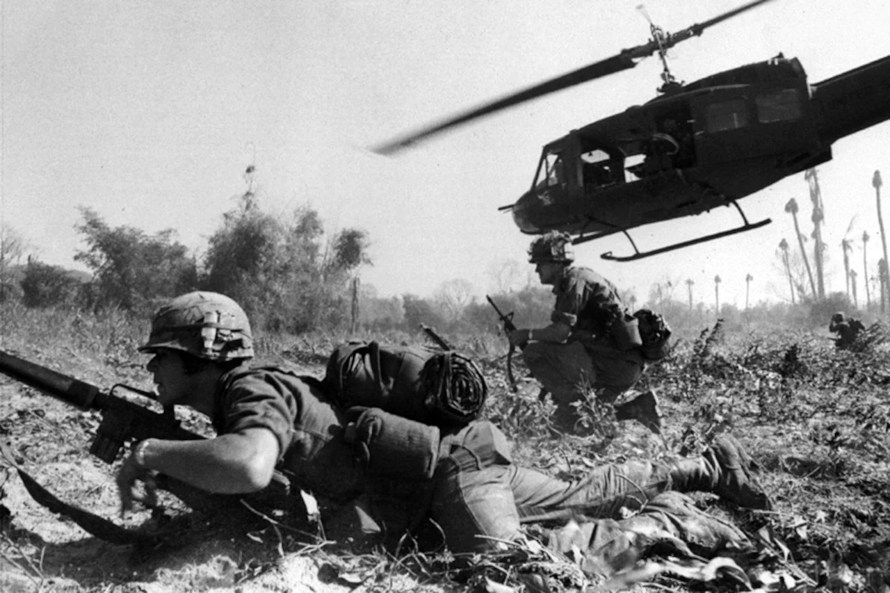 A helicopter flies close to ground troops in a field.