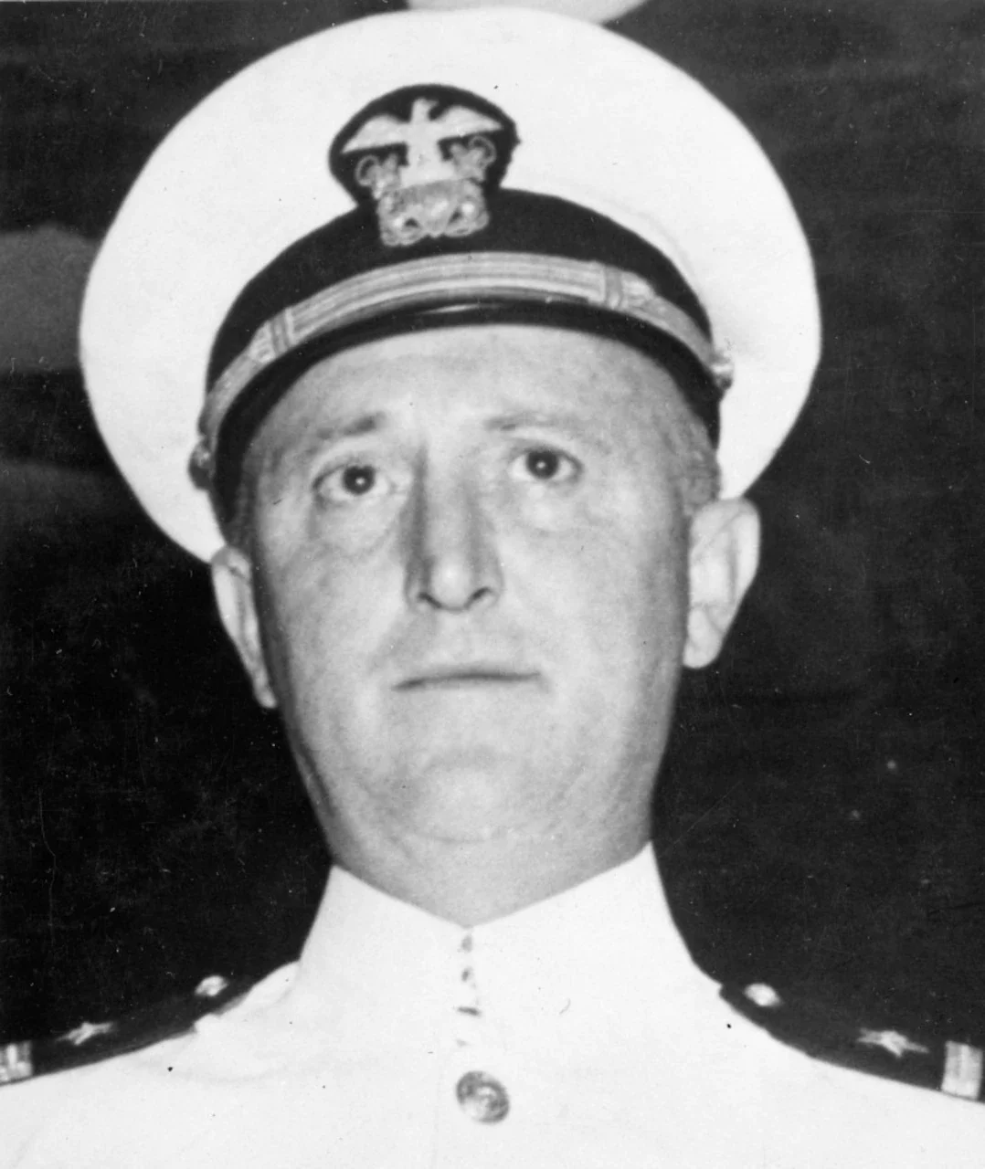 A man in a white dress uniform and white cap looks forward.