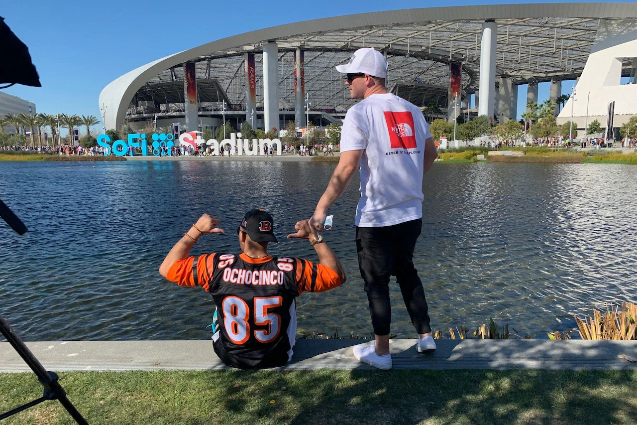A man sits on the ground pointing to his back. Another man stands beside him. They’re in front of a large pond across from a stadium.