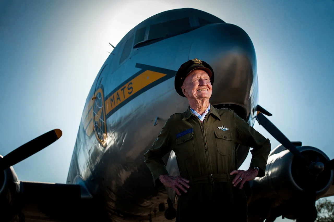 A smiling veteran in uniform stands in front of a shiny historic aircraft.