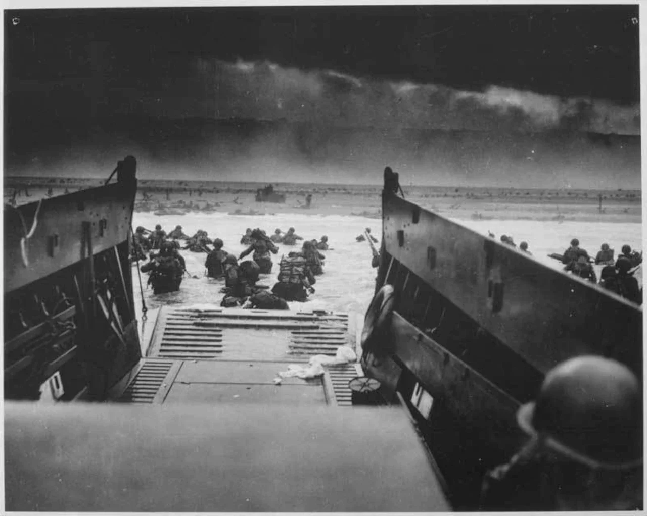 An empty boat looks over water filled with soldiers trudging to shore.
