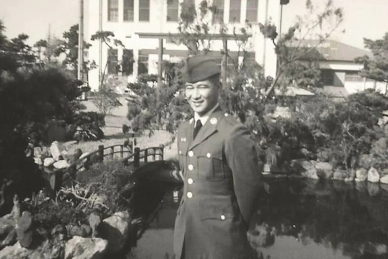 A soldier poses in a garden-like setting outside a building.