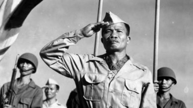 A soldier salutes while wearing a Medal of Honor, as fellow soldiers stand behind him outside and a flag waves above them.